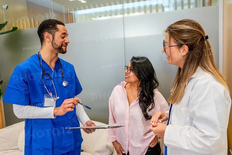 Patient consulting with a multi ethnic healthcare team members, a male nurse or doctor presenting information on a clipboard, and a female doctor listening attentively in a modern clinic setting