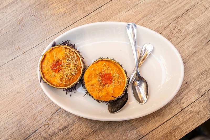Fresh sea urchin uni appetizer with salmon roe and shredded garnish on a white oval plate, two spoons at the side on a wooden table for a refined seafood bite