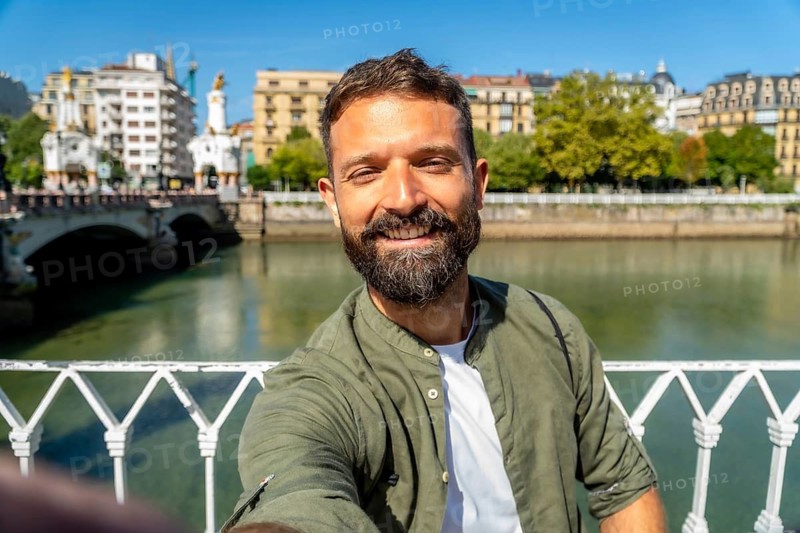 Smiling bearded man takes a selfie on maria cristina bridge over the urumea river, enjoying a sunny vacation day in san sebastian, spain, sightseeing and relaxed travel vibes