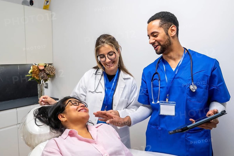 Diverse medical team internist and scrub clad clinician warmly engaging a smiling female patient during a modern clinic consultation, using tablet and records for collaborative care
