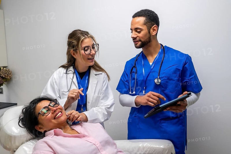 Healthcare staff, including a doctor and nurse, are discussing medical information with a smiling female patient, utilizing a digital tablet during a consultation in a clinic or hospital setting