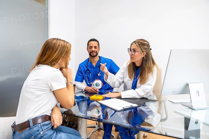 Medical team consisting of a female optometrist and male nurse consulting a female patient about eye health in a modern clinic office, using an anatomical eye model for explanation