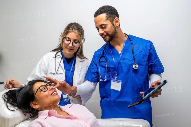 Medical professionals consulting a patient during a healthcare check up, with one doctor pointing to her nose while referencing information on a tablet in a modern clinic setting