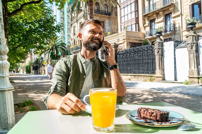 Smiling man with a beard sitting at an outdoor cafe table, having orange juice and a brownie while talking on a smartphone during a sunny day in the city