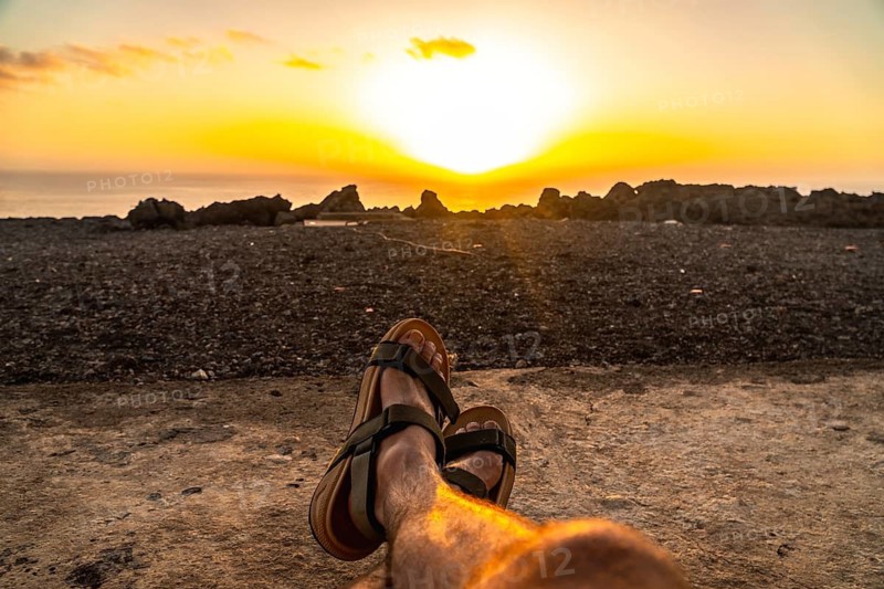 Person's feet wearing sandals relaxing on the beach sand, watching the golden orange sunset over the atlantic ocean and rocky coastline during a summer vacation in el hierro, canary islands, spain