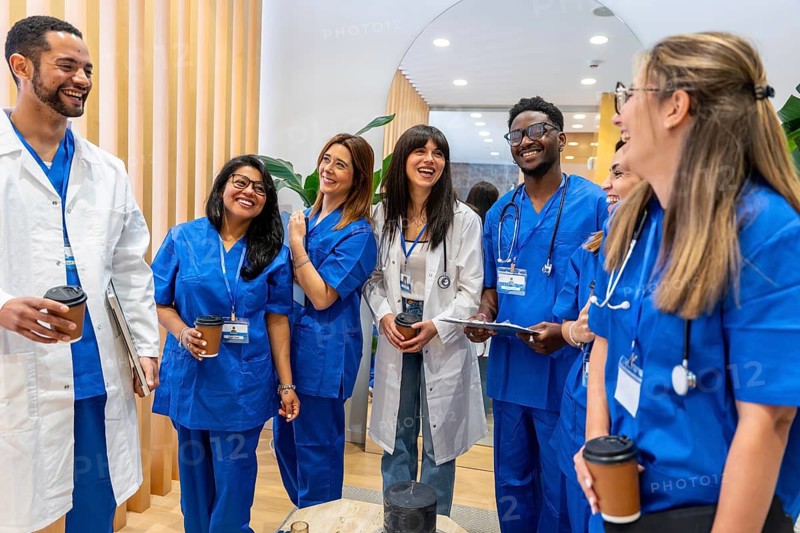 Diverse medical team in scrubs and lab coats sharing a joyful coffee break in a hospital corridor, laughing and chatting to celebrate teamwork, support and positive communication