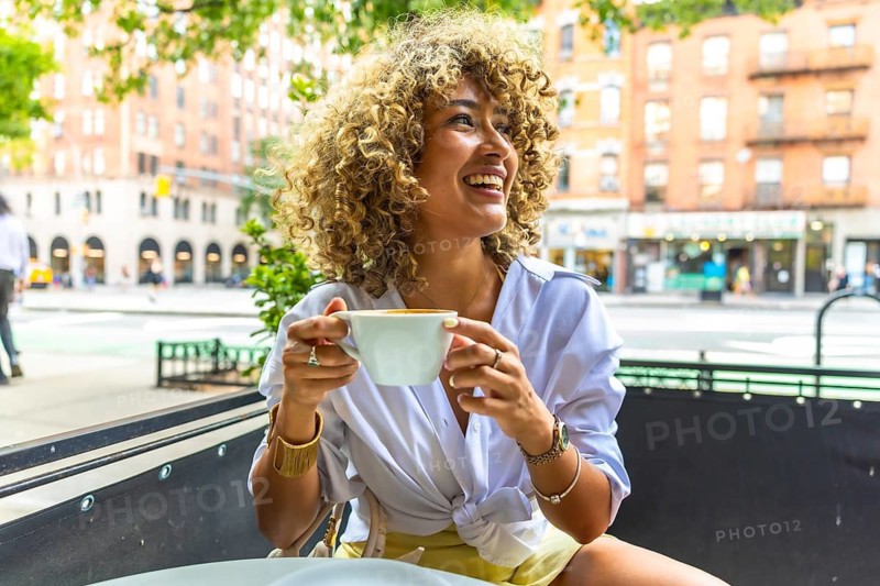 Woman with curly hair smiling and laughing, holding a coffee cup, sitting at an outdoor cafe on a vibrant street in manhattan, new york city