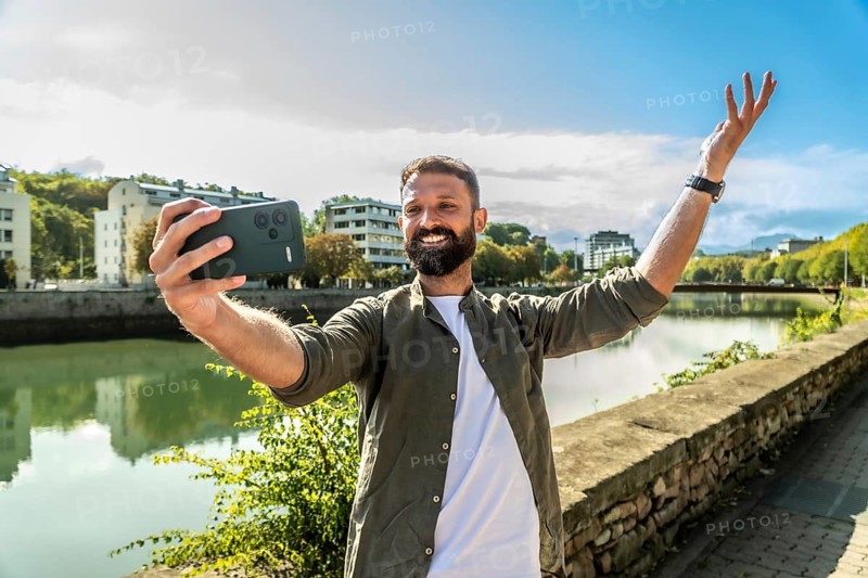 Happy man standing outdoors by a river and city buildings, smiling widely while recording a video or taking a selfie with a smartphone, celebrating his travel moment