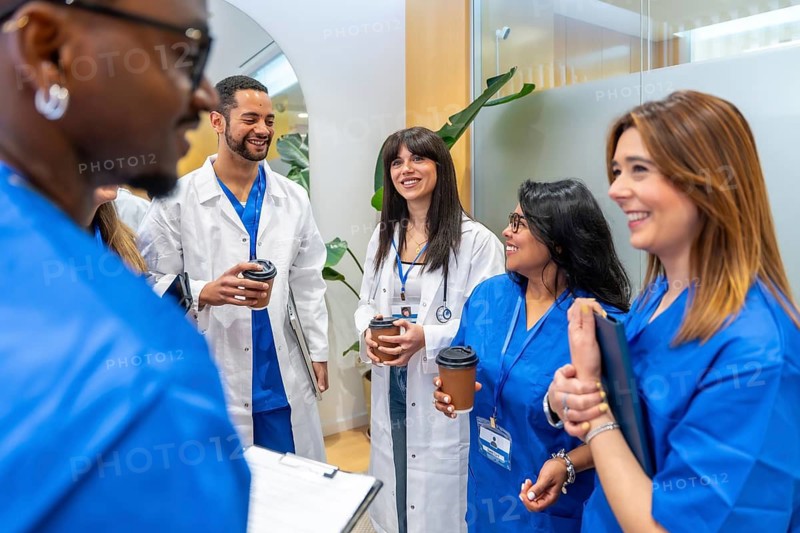 Medical staff, including doctors and nurses, smiling and discussing while holding coffee cups during a break in a modern hospital or clinic setting, conveying teamwork and collaboration