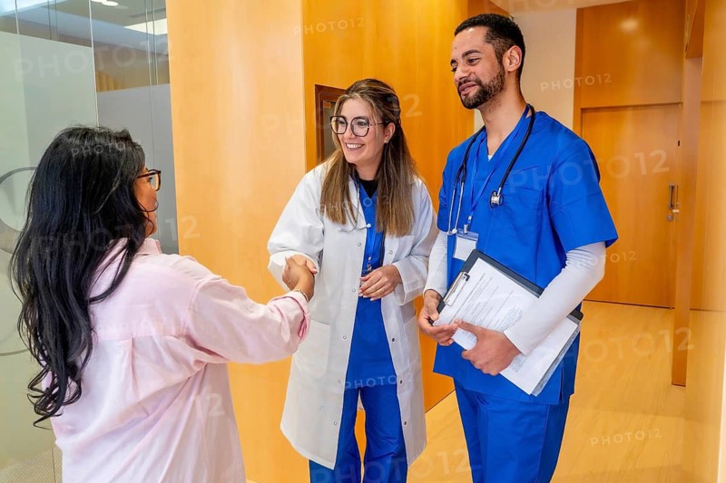 Female doctor in lab coat and male nurse in scrubs warmly greet a female patient with a handshake in a modern hospital corridor, highlighting teamwork and compassionate care