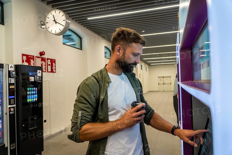 Man holding a coffee cup, using a touch screen interface on an automated vending machine in a bright, contemporary interior space, symbolizing modern convenience and urban lifestyle