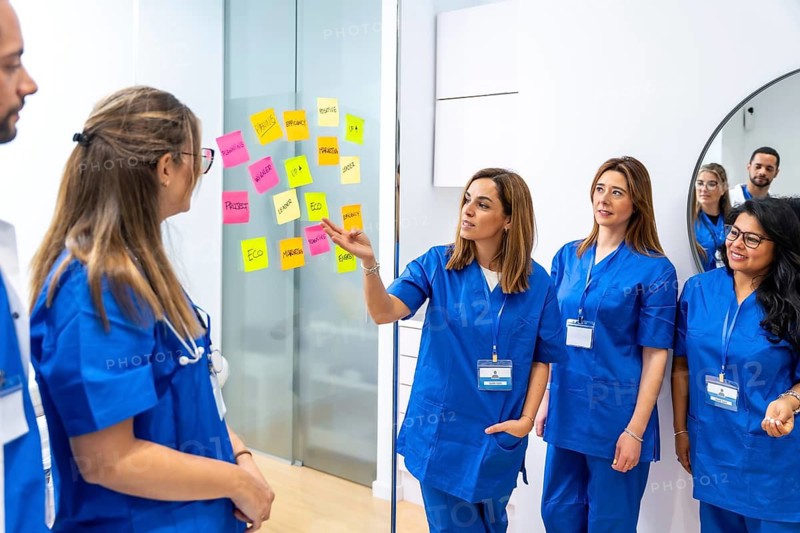Medical team in blue scrubs gathered at a glass board with colorful sticky notes, collaboratively brainstorming and planning improvements for patient care and hospital workflows