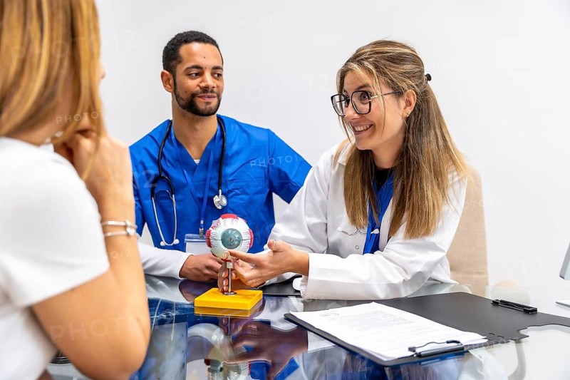 Medical team consisting of two clinicians educating a female patient about eye anatomy and ophthalmology concepts using a detailed anatomical model during a clinic visit