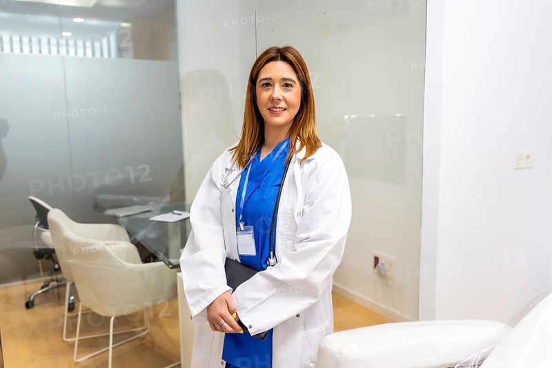 Female doctor in a white lab coat and blue scrubs smiling confidently, holding a tablet while standing in a contemporary medical office or hospital setting