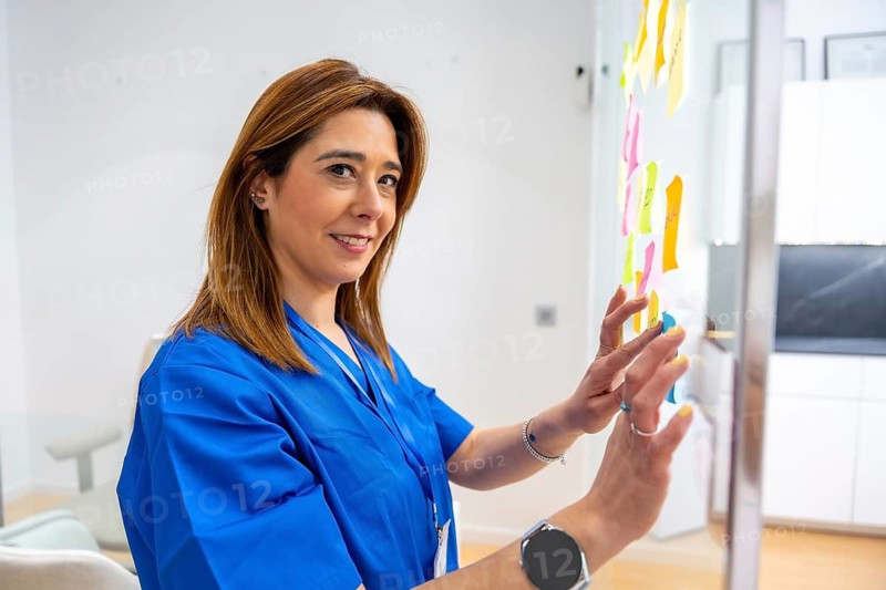 Healthcare professional wearing blue scrubs smiling while organizing colorful sticky notes on a glass whiteboard, planning medical concepts and strategies within a modern medical center environment