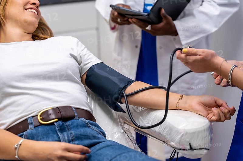 Patient lying on a medical chair, smiling while a healthcare professional measures her blood pressure in a clinic, with another doctor reviewing patient data on a tablet in the background