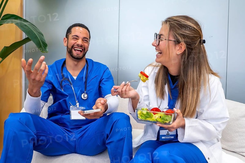 Two cheerful medical professionals enjoy a moment of laughter and camaraderie while sharing a healthy meal during their break at a hospital or clinic, promoting well being and teamwork