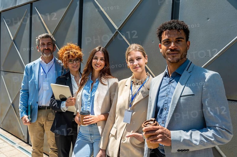 Diverse group of male and female business professionals standing together, smiling and holding drinks and a tablet, showcasing teamwork, success, and community in an outdoor office setting