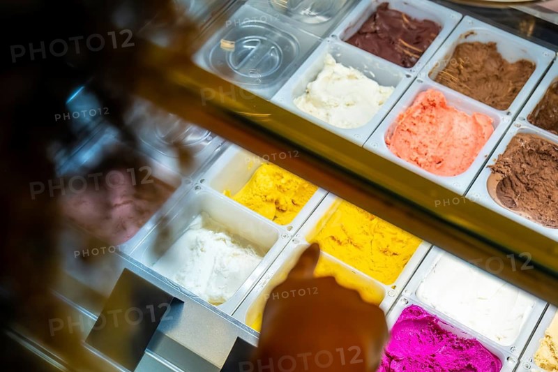 Customer hand pointing at brightly colored gelato and sorbet flavors in a refrigerated display case, highlighting variety and delicious dessert options in a sweet shop