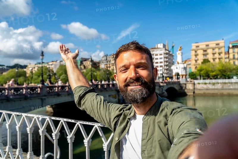 Happy bearded man smiling and taking a selfie portrait during a sunny day trip, enjoying travel and tourism in san sebastian with the maria cristina bridge in the background