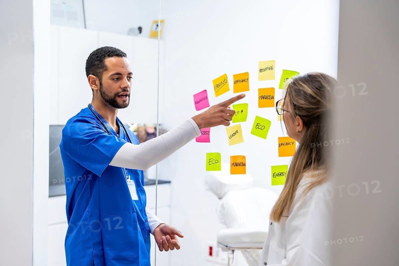 Healthcare team in scrubs and white coats brainstorm and prioritize ideas on a glass board with colorful sticky notes, collaborating on strategy and problem solving in a modern clinic