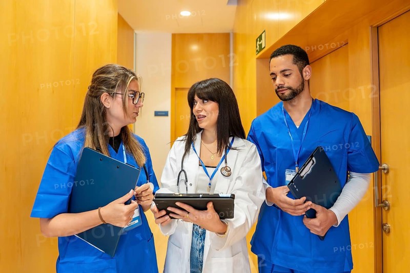 Healthcare professionals wearing scrubs and lab coat collaborating, discussing patient data on a digital tablet and clipboard while walking in a hospital or clinic hallway