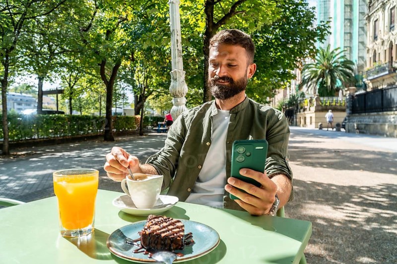 Bearded man relaxing at a sunny outdoor cafe, enjoying coffee, orange juice, and chocolate cake while using a smartphone, connecting with technology in a city environment