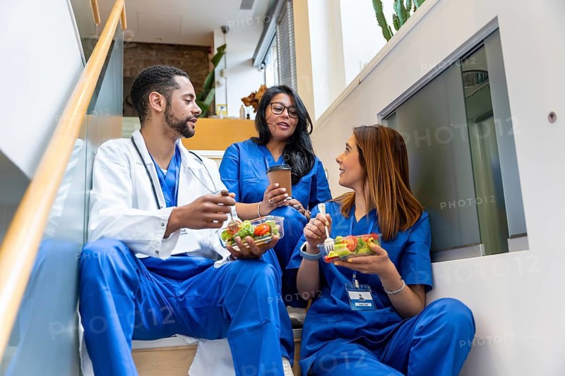 Multiracial medical team colleagues sitting on a staircase, enjoying their healthy lunch during a break, and engaging in conversation about their work in the modern hospital environment