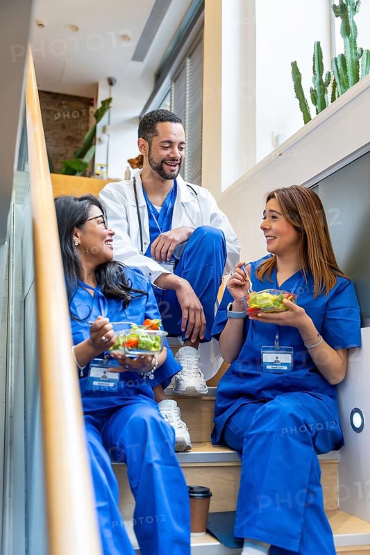 Diverse medical team in scrubs and a lab coat sitting on a staircase in a modern clinic, smiling and chatting while enjoying salads during a relaxed lunch break