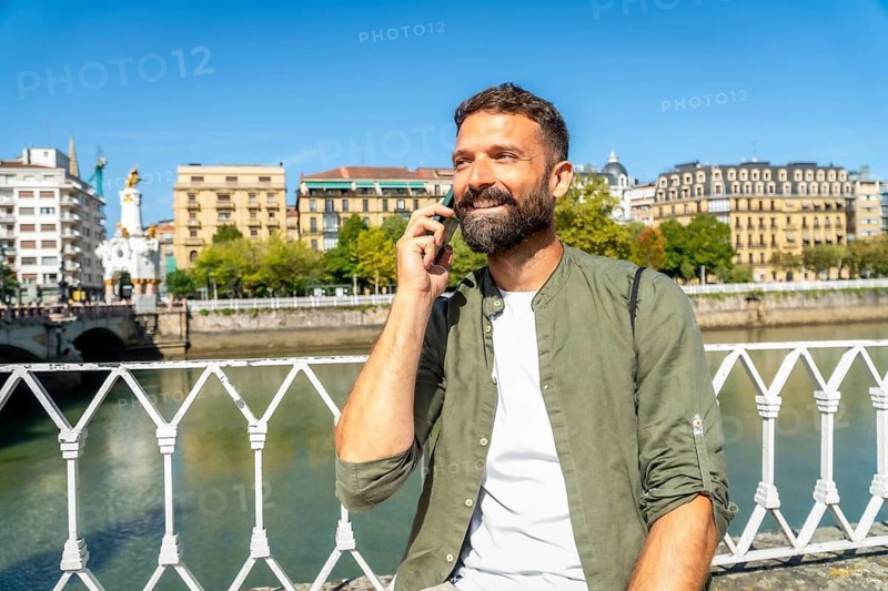 Smiling man talking on his smartphone on maria cristina bridge, enjoying sunny views over the urumea river and historic buildings in san sebastian, spain during summer tourism