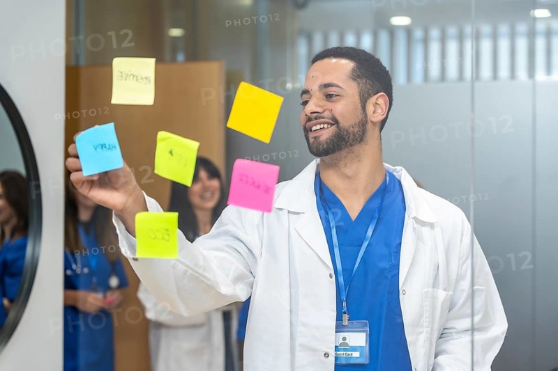 Doctor in medical uniform and lab coat brainstorming and organizing ideas on a glass wall with colorful sticky notes, colleagues smiling in background, focusing on healthcare concepts
