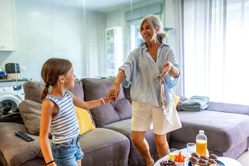 Grandmother holding granddaughter's hand, looking joyful while dancing in a bright living room at home, sharing a happy intergenerational moment expressing love and connection