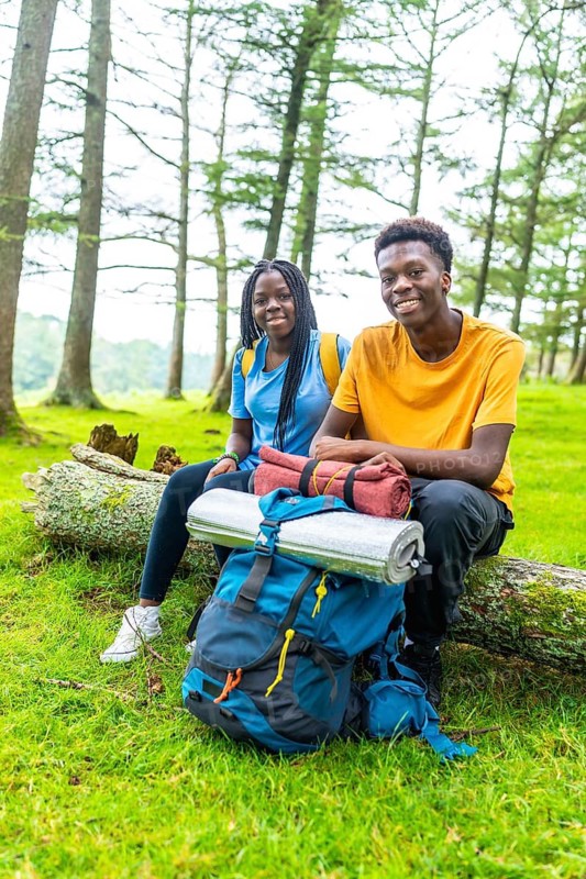 Vertical portrait of two young male and female african friends smiling while sitting on a log in a beauty forest