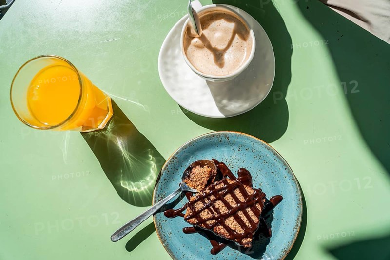 Breakfast spread on a green outdoor table with coffee, orange juice and cake, bright harsh sunlight casts strong shadows for a casual, vibrant summer morning mood