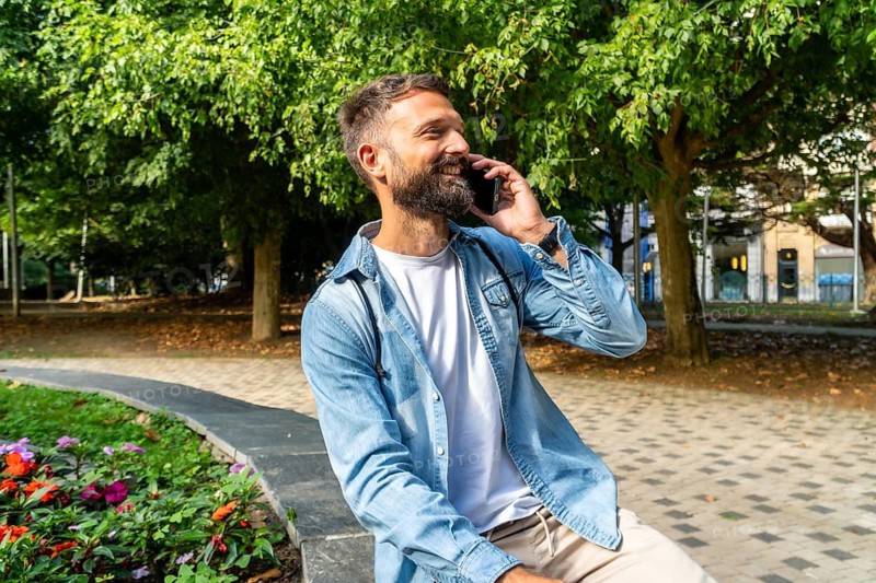 Bearded man sitting in a city park, smiling and chatting on his smartphone, enjoying a relaxed summer moment outdoors surrounded by trees and urban greenery