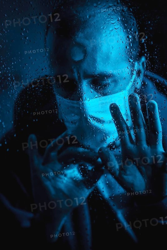 A young man wanting to leave home in the covid 19 quarantine one rainy night with a mask looking out the window, with blue ambient light