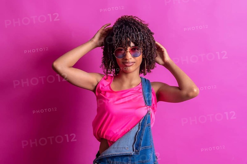 Fashionable young woman with curly hair, wearing a pink top and denim overalls, adjusts her sunglasses against a vibrant pink backdrop