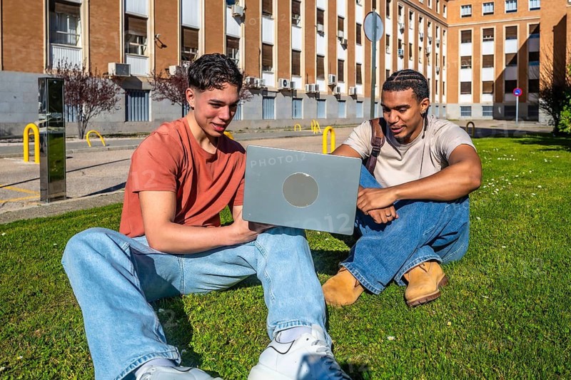 Two diverse young men sitting on a university lawn, collaborating on a laptop, smiling and studying together outdoors in sunny spring weather, relaxed and focused on a project