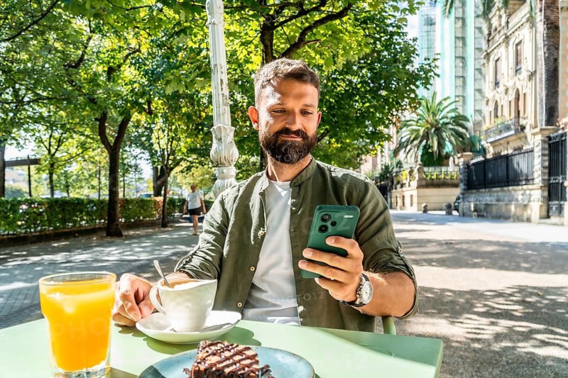 Bearded young man in a casual shirt enjoys breakfast at an outdoor cafe, checking his smartphone amid coffee, juice and a pastry with green trees and city buildings nearby on a sunny day