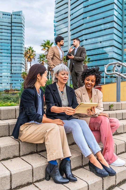 Vertical photo of three multi-ethnic businesswomen sitting in outdoor urban stairs working with digital tablet
