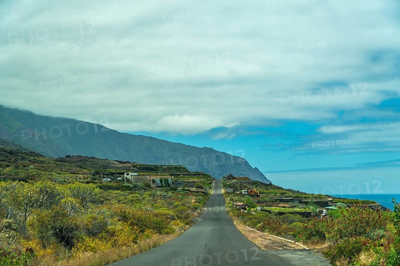 Winding asphalt road navigating through the lush volcanic landscape of el hierro island, with distant mountains, coastal vegetation, and the vast atlantic ocean under a cloudy sky