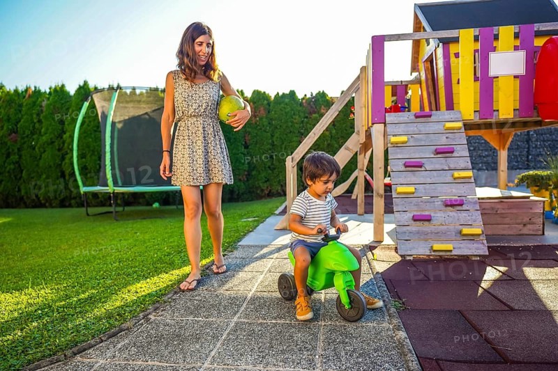 Mother holding ball walking and watching her son riding toy motorbike in backyard on summer vacation