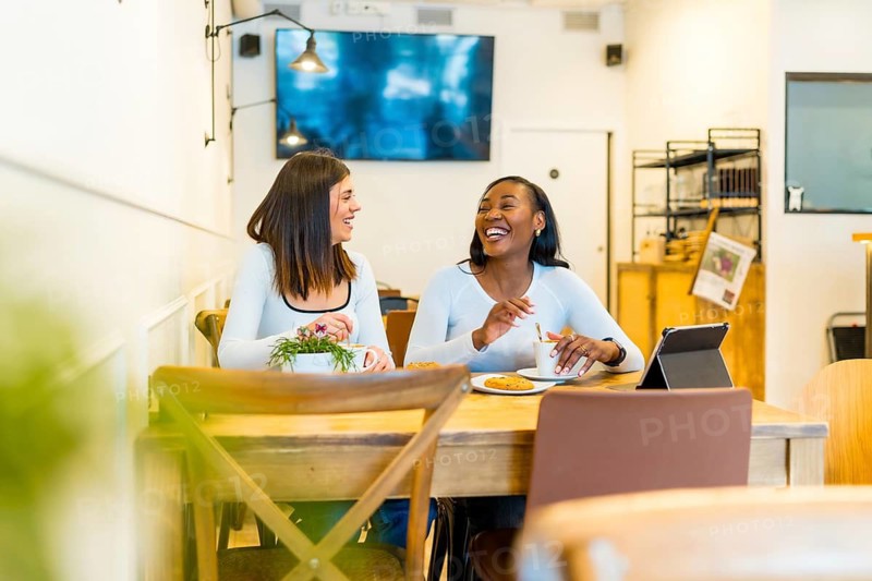 Two young female friends enjoying a coffee break, laughing and talking together at a table in a modern cafe