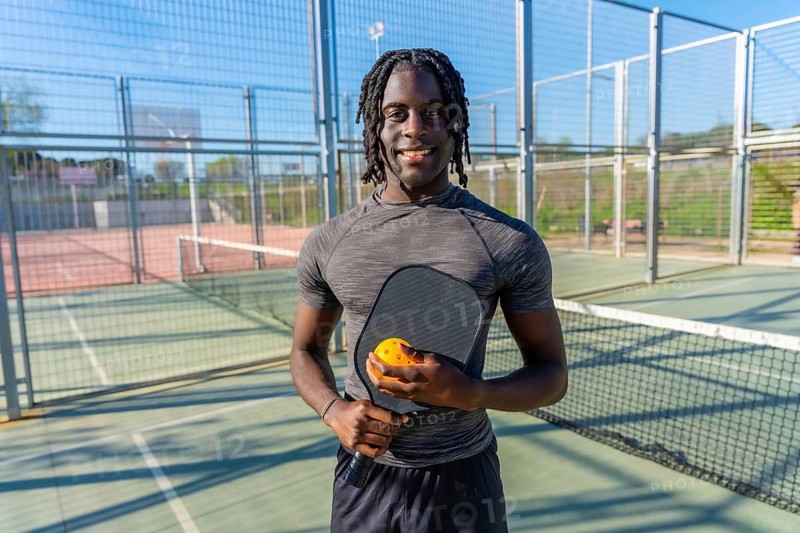 Young athletic black man standing on an outdoor pickleball court, holding a paddle and ball, smiling and looking at the camera, promoting healthy active lifestyle and sport