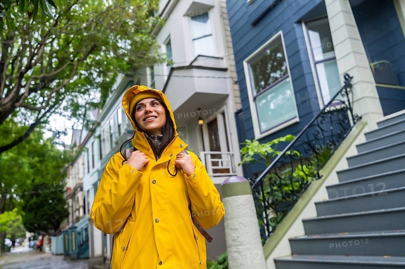 Young woman wearing a yellow raincoat and backpack, looking up while walking along a street lined with traditional victorian houses in san francisco, discovering the urban architecture