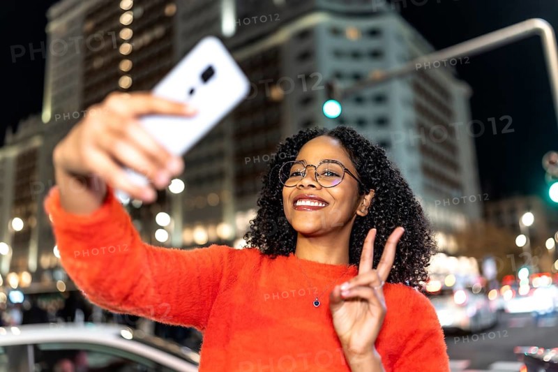 Smiling young woman with curly hair and glasses taking a selfie with smartphone and making peace sign in a city at night