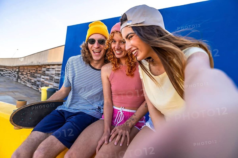 Three cheerful young friends are sitting on a rooftop, taking a selfie with a skateboard nearby