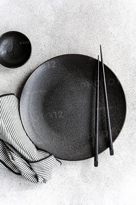 A minimalist black and white table setting featuring a textured dark plate, matching bowl, pair of chopsticks, and a striped cloth on a light background