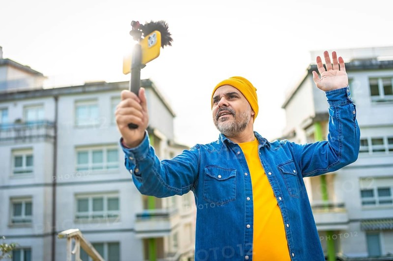 Low angle view portrait of a mature male vlogger waving at audience streaming in the city using smartphone and microphone