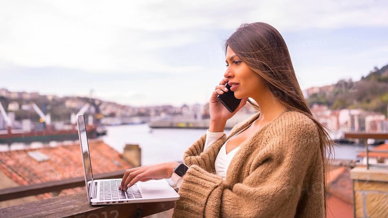A young Latin woman with a white t-shirt and a brown wool sweater, teleworking on the terrace of a house by the sea, talking on the phone